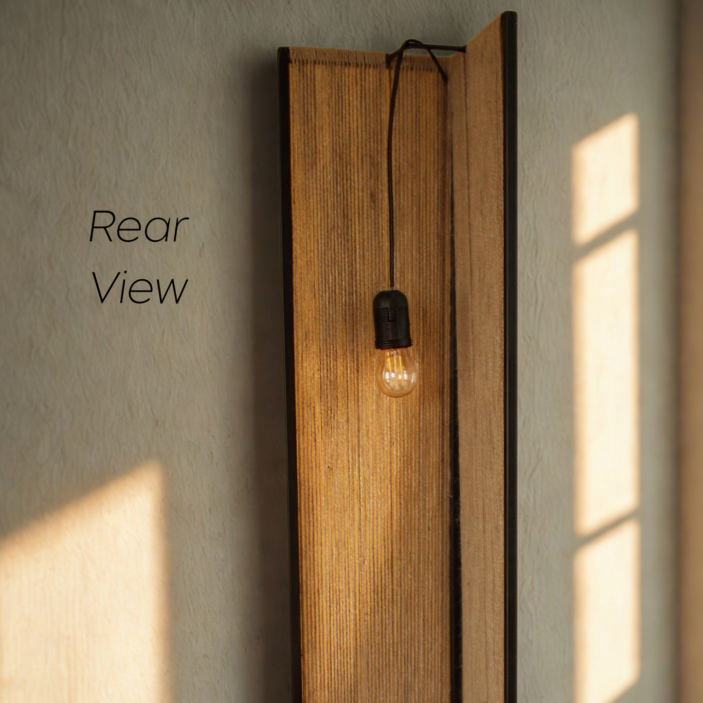 View of light fixture with a tall rectangular twine panel backplate and a single black cord holding an exposed-arc light bulb, shown against a textured beige wall with sunlight casting shadows. The text 'Rear View' appears on the left.