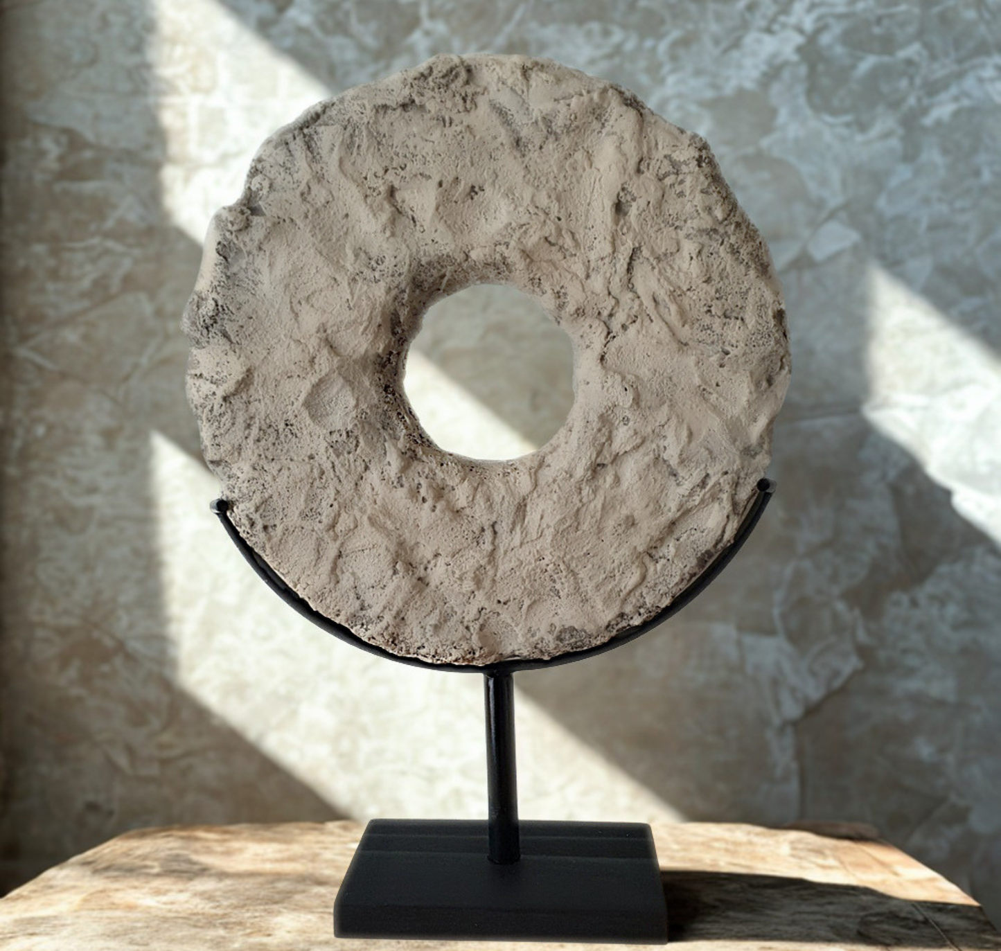 A textured circular stone or concrete ring sculpture with a central hole, displayed on a black metal stand against a stone-patterned wall with dramatic sunlight and shadows.