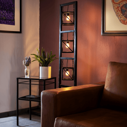 Modern black metal floor lamp with three pendant lights, placed on a tiled floor next to a brown sofa with a purple wall in the background, with a decorative item on the side table.
