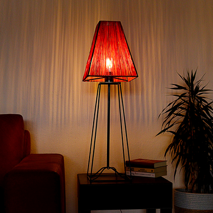 A table lamp with a black base and reddish twine lampshade, illuminated, placed on a wooden table next to a brown sofa in a dimly lit room with warm light streaks