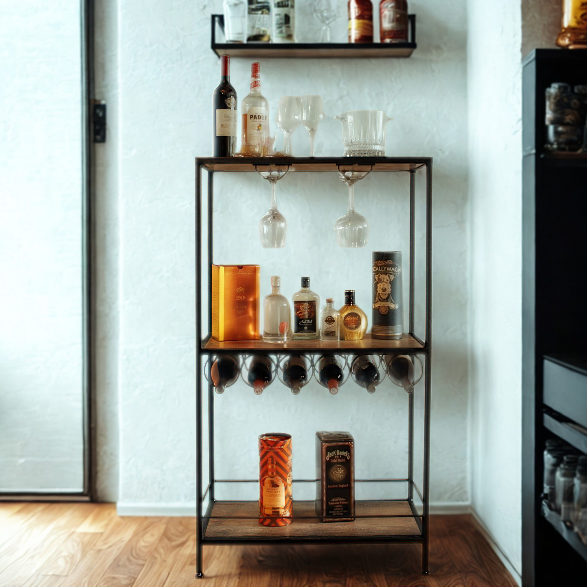 A tall black metal bar cart with three wooden shelves, styled with various liquor bottles, glasses, and decorative items, set against a white wall on a wooden floor.