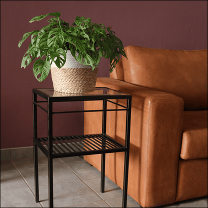 A stylish black metal and glass table with two shelves, known as the Ardhi Manii Table, featuring a woven basket-top plant, placed on a brown leather chair with a purple wall in the background.