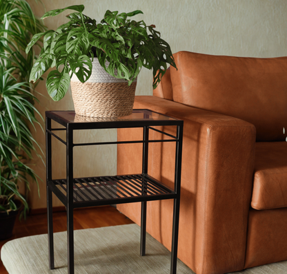 A black metal two-tier side table with a glass top, styled in a living room next to a brown leather sofa with a potted green plant on top.
