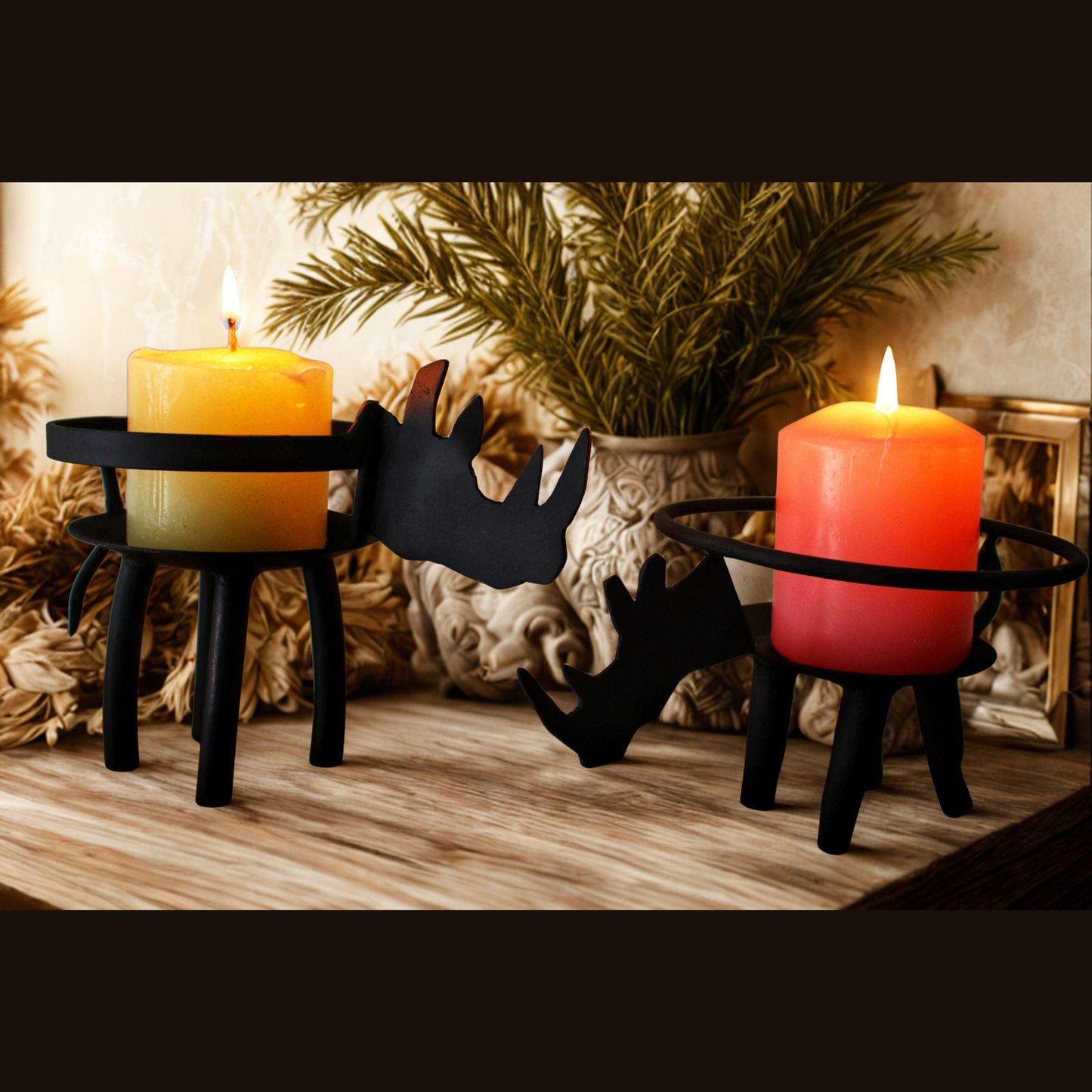 Two black rhino-shaped candle holders on a wooden surface, each holding a lit pillar candle, with a potted plant and decorative background elements behind them.