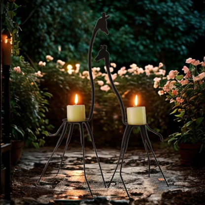Two black metal giraffe-shaped candle holders with lit candles, placed on a stone path in a garden at night with blurred flowers and foliage in the background.