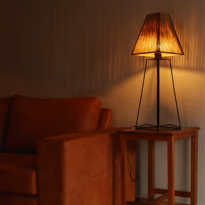A table lamp with a black base and natural twine beige lampshade, illuminated, placed on a wooden table next to a brown sofa in a dimly lit room.
