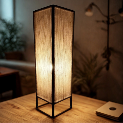 A modern geometric floor lamp with a black metal frame and textured beige woven shade, glowing warmly on a wooden table in a cozy room setting.