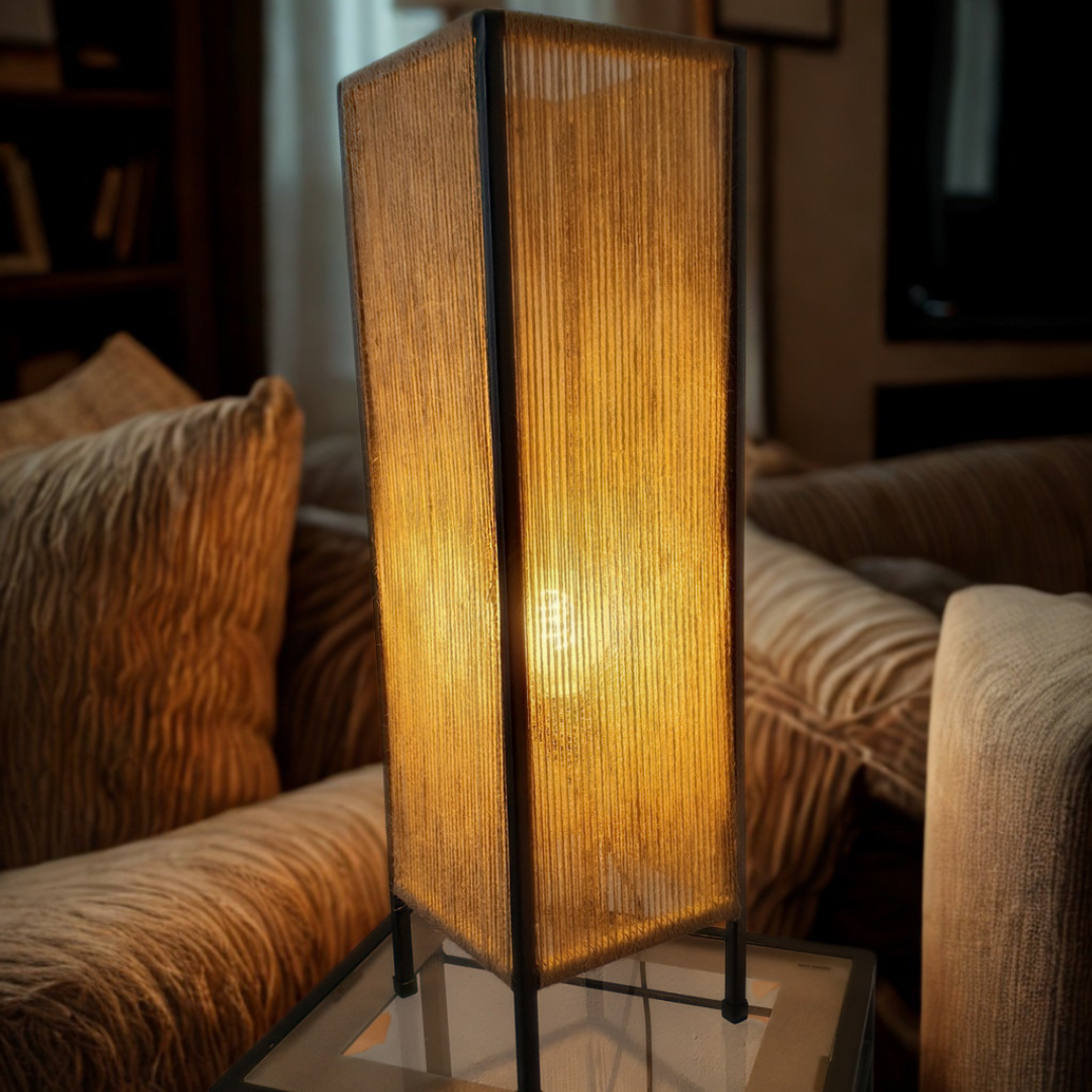 A modern rectangular floor lamp with a glowing warm yellow light inside a textured beige woven shade, supported by a slim black metal frame, set on a glass-topped side table in a cozy living room next to a striped sofa.