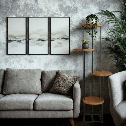 A styled living room corner featuring a light gray fabric sofa with matching cushions, a three-panel abstract wall art set, a tall black metal floor shelf with four wooden tiers holding plants, and a textured gray concrete wall background.