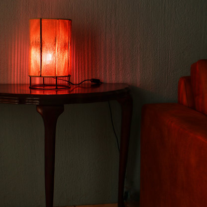 A table lamp with a rose pink cotton shade, emitting a warm light, placed on a side table next to a red armchair