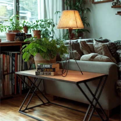 A modern coffee table in a cozy living room setting, featuring a light wood top and a black metal X-frame base. A potted green plant and a lit floor lamp sit on the table.
