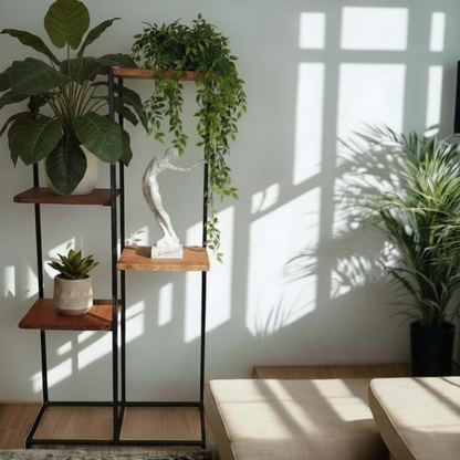 A tall black metal shelving unit with four wooden shelves stands in a sunlit room, styled with potted green plants, a small succulent, and a white figurative sculpture. The shelves are set against a white wall with strong window shadow patterns, and a beige sofa is partially visible in the foreground.