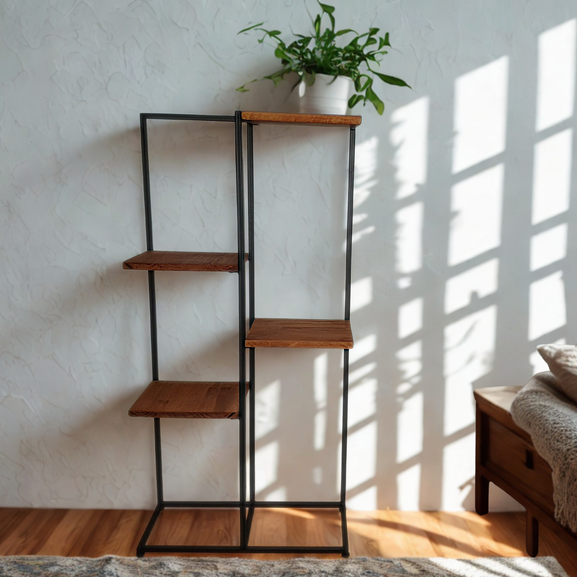 A tall black metal shelving unit with five wooden shelves stands against a textured white wall in a sunlit room. A potted green plant sits on the top shelf, and a wooden bench is partially visible on the right.