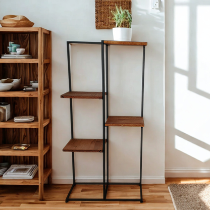 A tall black metal ladder-style shelving unit with four warm brown wooden shelves stands in a bright room next to a larger wooden bookcase. A small potted plant sits on the top shelf, and sunlight streams in from the right side of the room.