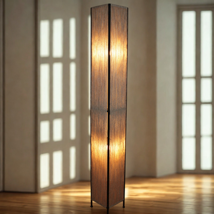 A tall rectangular floor lamp with a warm glowing light inside and a dark natural twine shade, standing on a wooden floor in a softly lit room with window panes in the background.