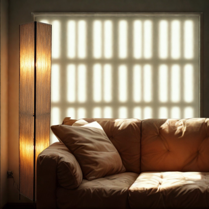 A warm, modern interior scene featuring a tall, slender metal and twine floor lamp glowing on the left, casting vertical shadows across a tufted brown sofa with cushions. The background shows a window with blinds.