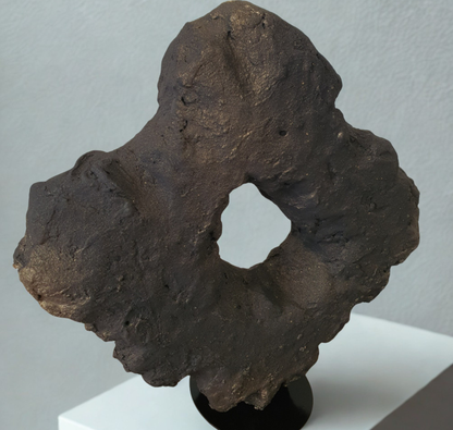 A dark black and light gold textured stone or rock specimen with a central hole, displayed on a black stand atop a white pedestal against a light gray wall.