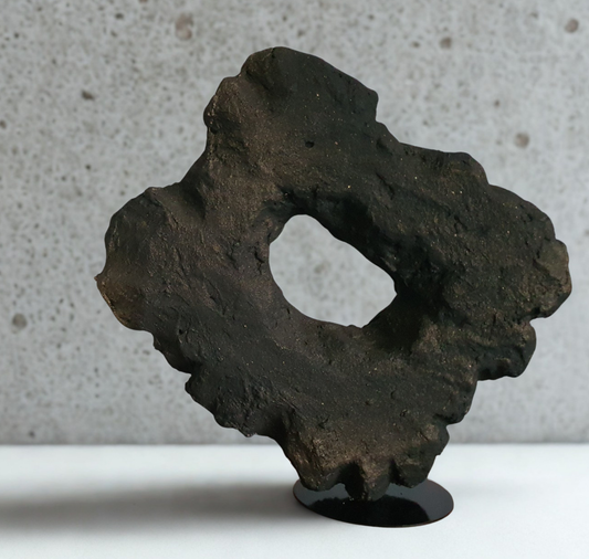 A dark, textured abstract stone sculpture with a central hole, displayed on a small black round base against a light gray speckled wall and white surface.
