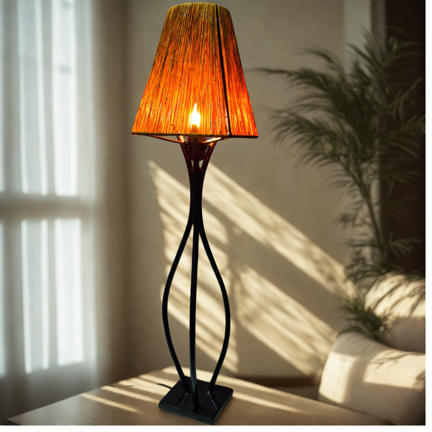 A table lamp with a warm orange textured shade and a black sculptural base, shown lit in a softly lit room with sunlight and shadows.