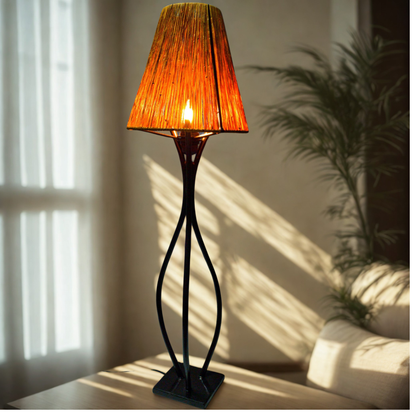 A table lamp with a warm orange textured shade and a black sculptural base, shown lit in a softly lit room with sunlight and shadows.